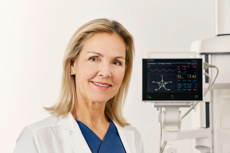 A woman in a lab coat stands smiling beside medical equipment with digital displays in a clinical setting.