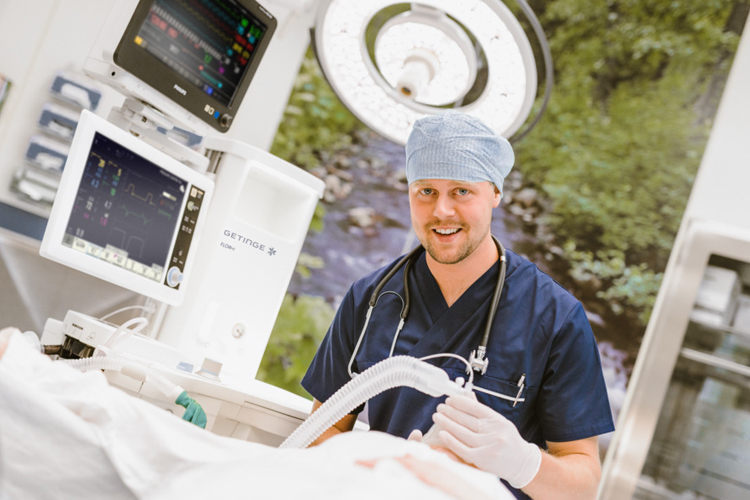 Smiling healthcare professional in scrubs with a stethoscope, standing next to medical equipment in a hospital.