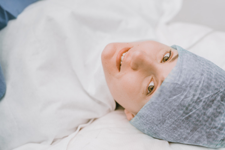 Smiling person lying down, wearing a gray surgical cap and white gown.