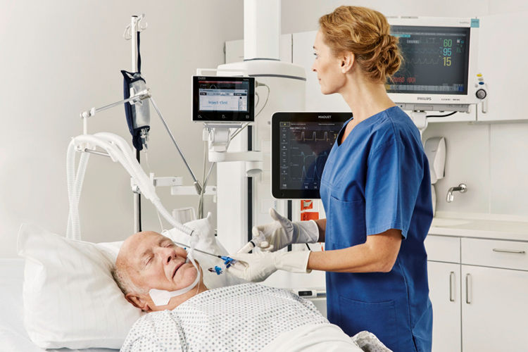 Healthcare worker in scrubs cares for an elderly patient using a ventilator in a hospital room.