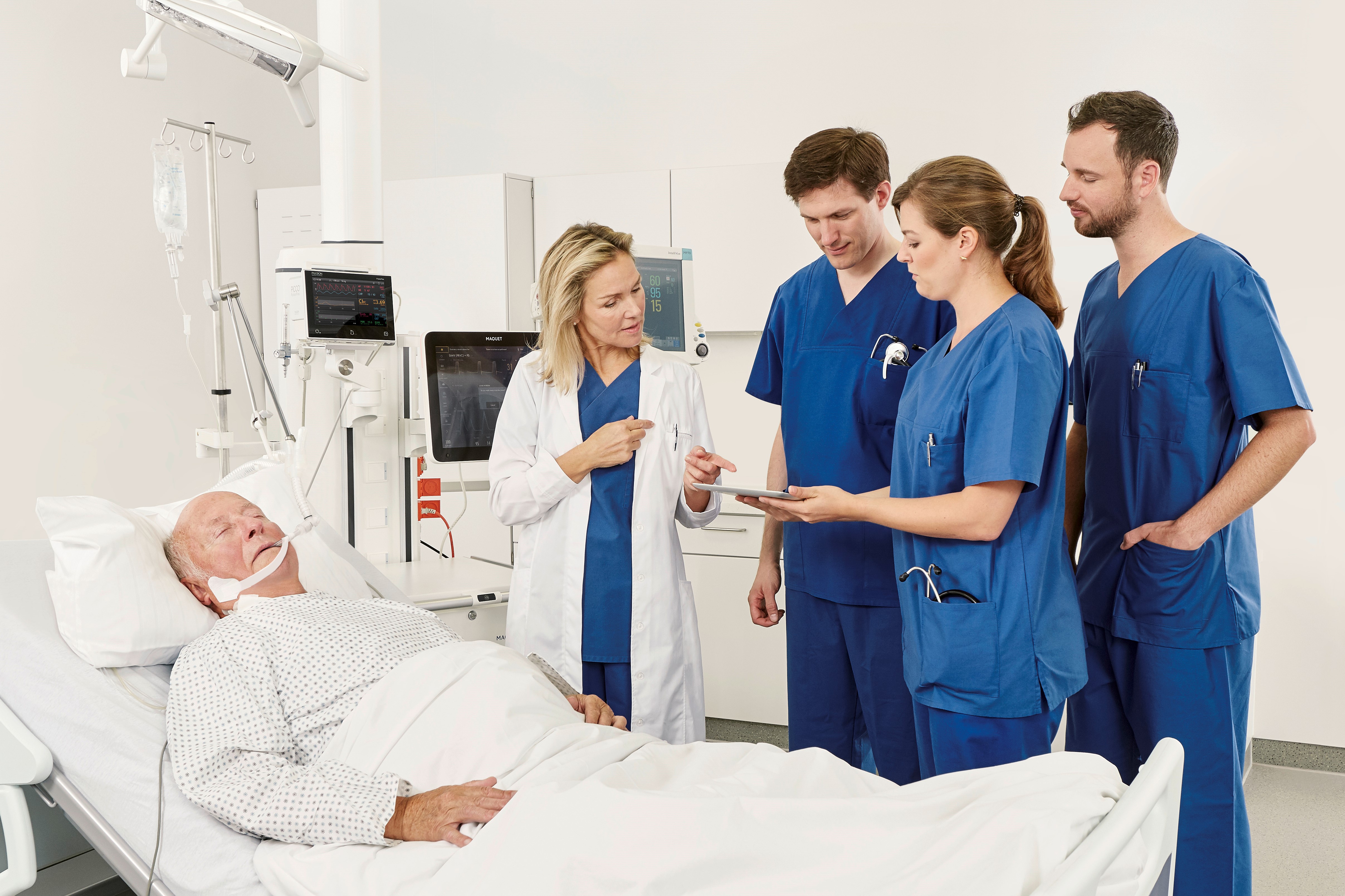 Medical team in blue scrubs and a doctor in a white coat discuss near a patient lying in a hospital bed.