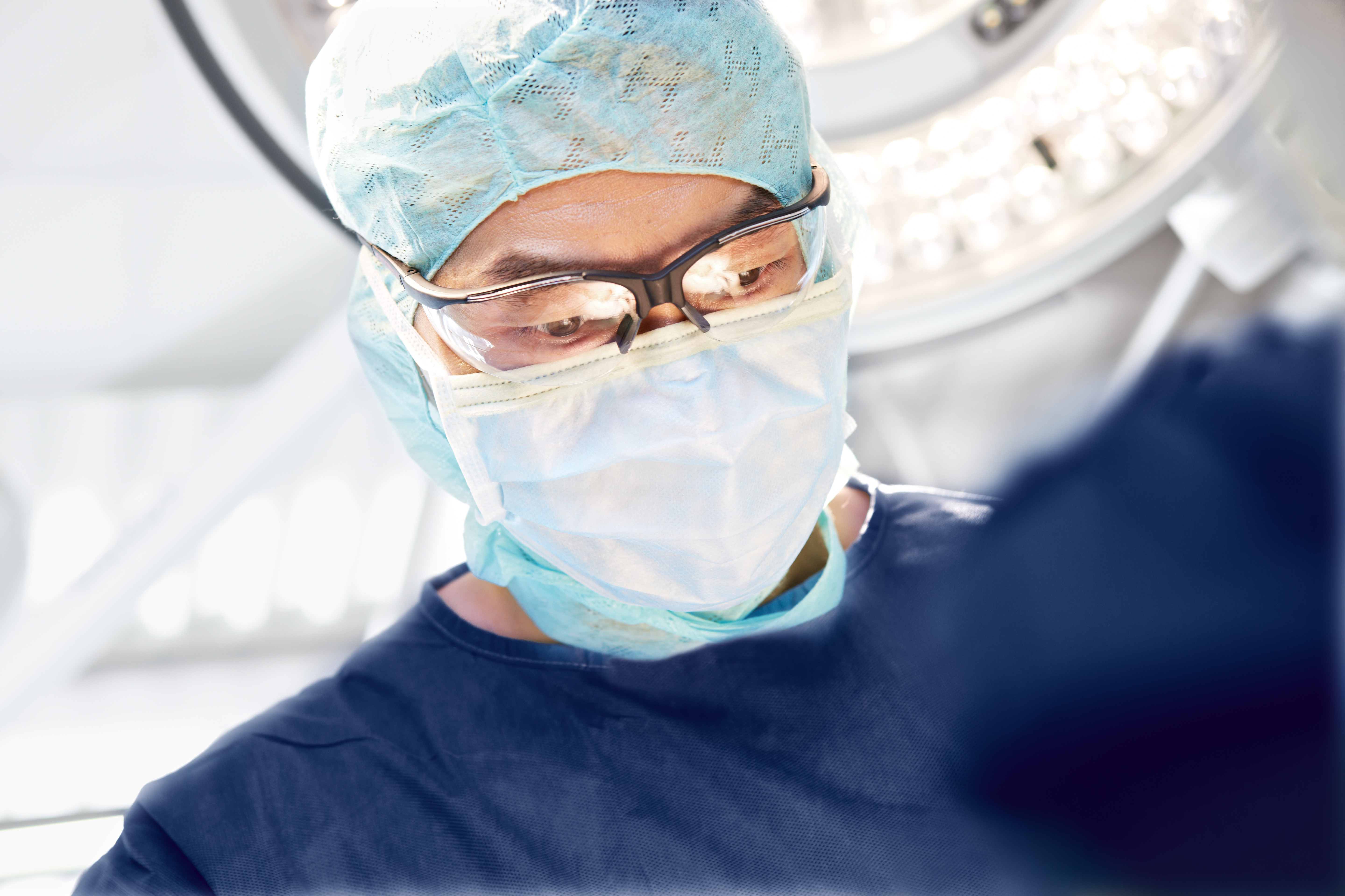 Surgeon in mask and cap focused on procedure under bright surgical lights.