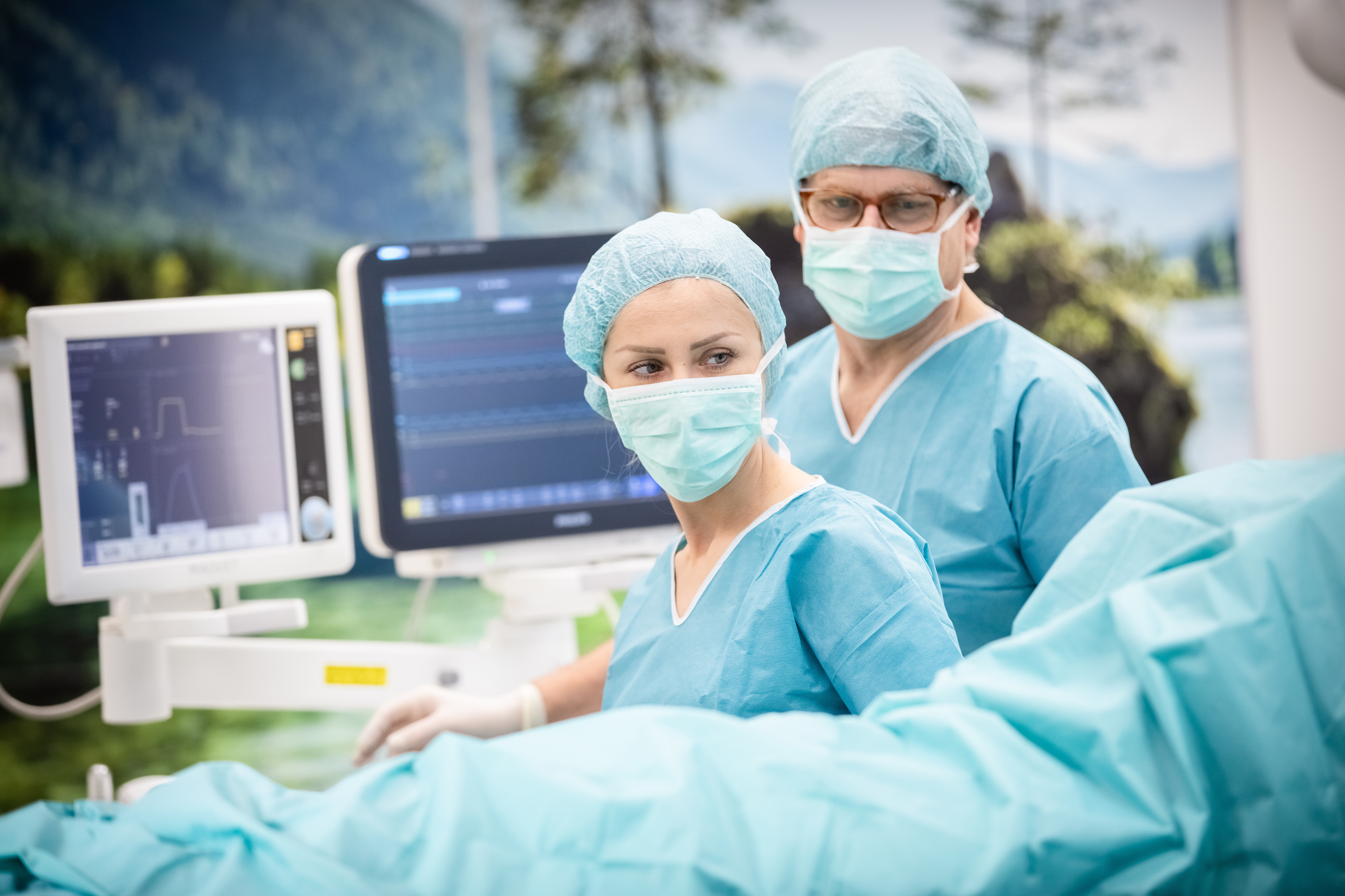 Two surgeons in scrubs and masks work in an operating room with medical monitors in the background.