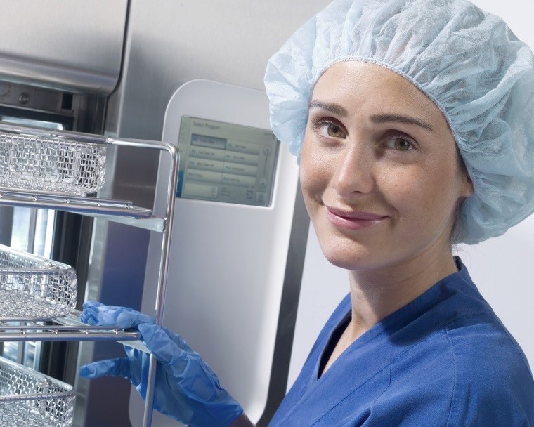 Person in blue scrubs and hairnet, wearing gloves, handling metal trays near medical equipment, smiling at the camera.