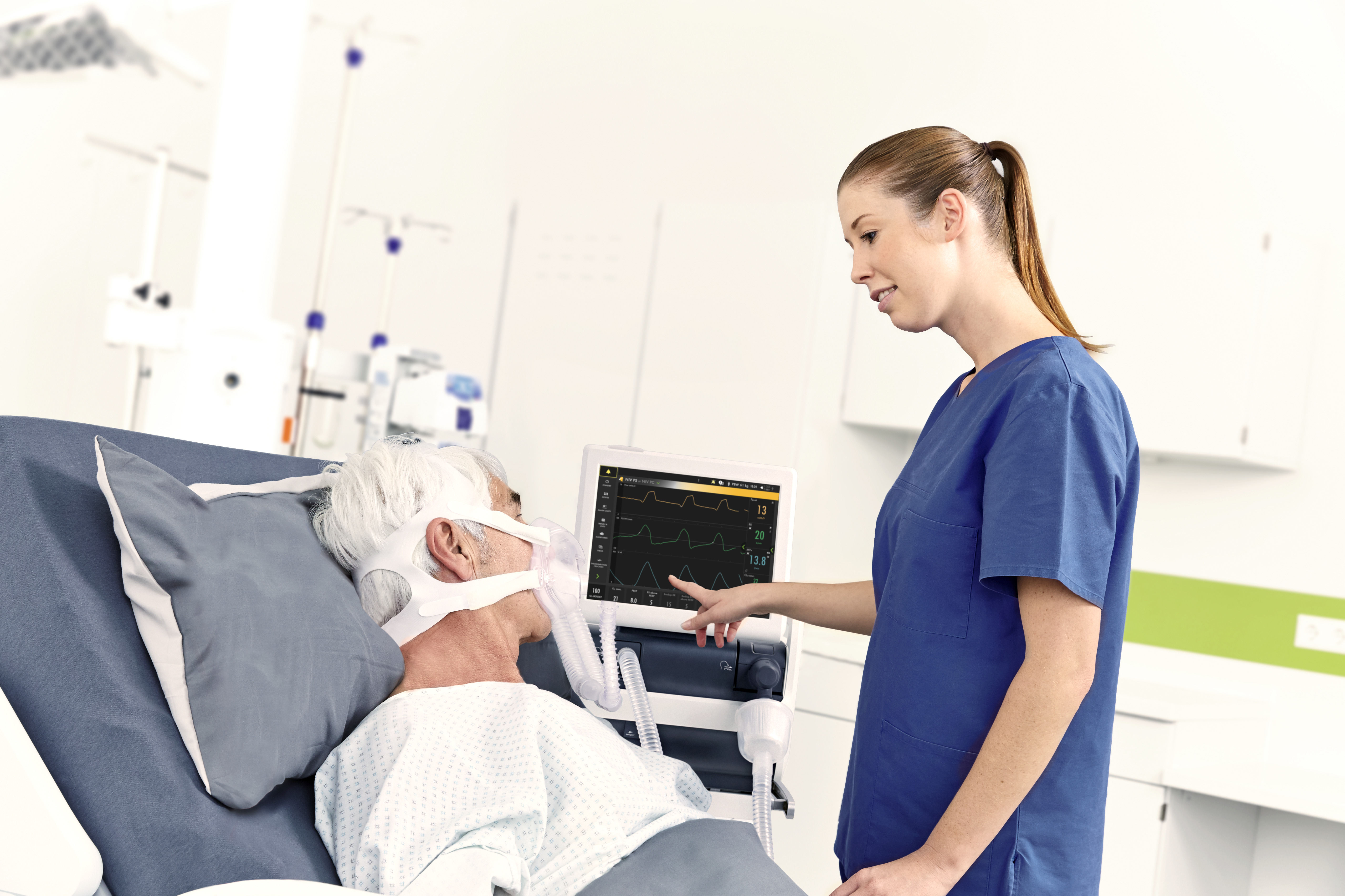 Healthcare worker in blue scrubs checks monitor for elderly patient with oxygen mask in a hospital bed.