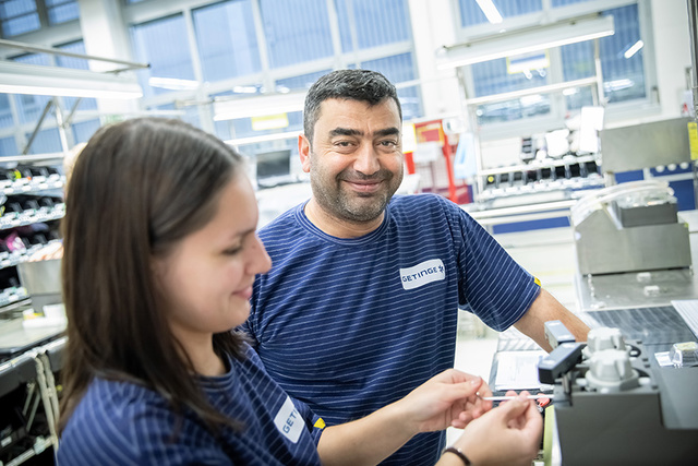 Zwei Personen in gestreiften Uniformen in einer Fabrik. Einer lächelt und der andere hält Gerätekomponenten.