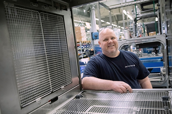 Ein lächelnder Mann steht in einer Fabrik neben Industrieanlagen. Im Vordergrund sind Metallgitter und Regale zu sehen.