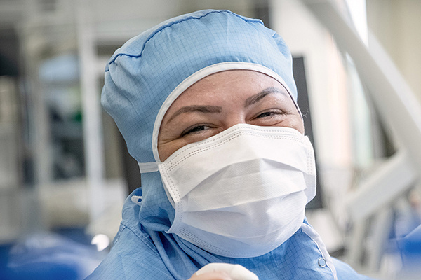 Person im blauen Arztkittel und mit Maske lächelt in einem Krankenhaus oder Labor.