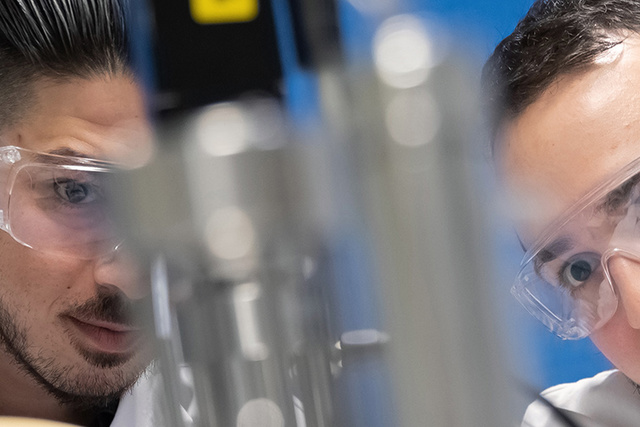 Two men in lab coats and safety goggles focus intently on a piece of laboratory equipment.