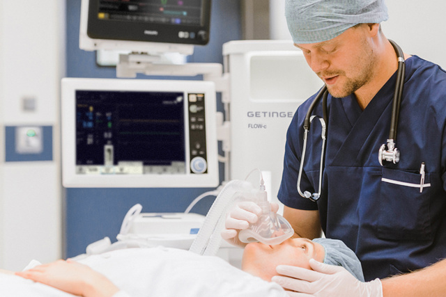 Een zorgverlener houdt een zuurstofmasker op het gezicht van een patiënt, met een Flow-c anesthesietoestel op de achtergrond.