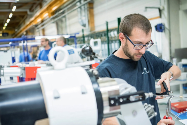 Een man met een bril werkt aan machines in een drukke industriële werkplaats.