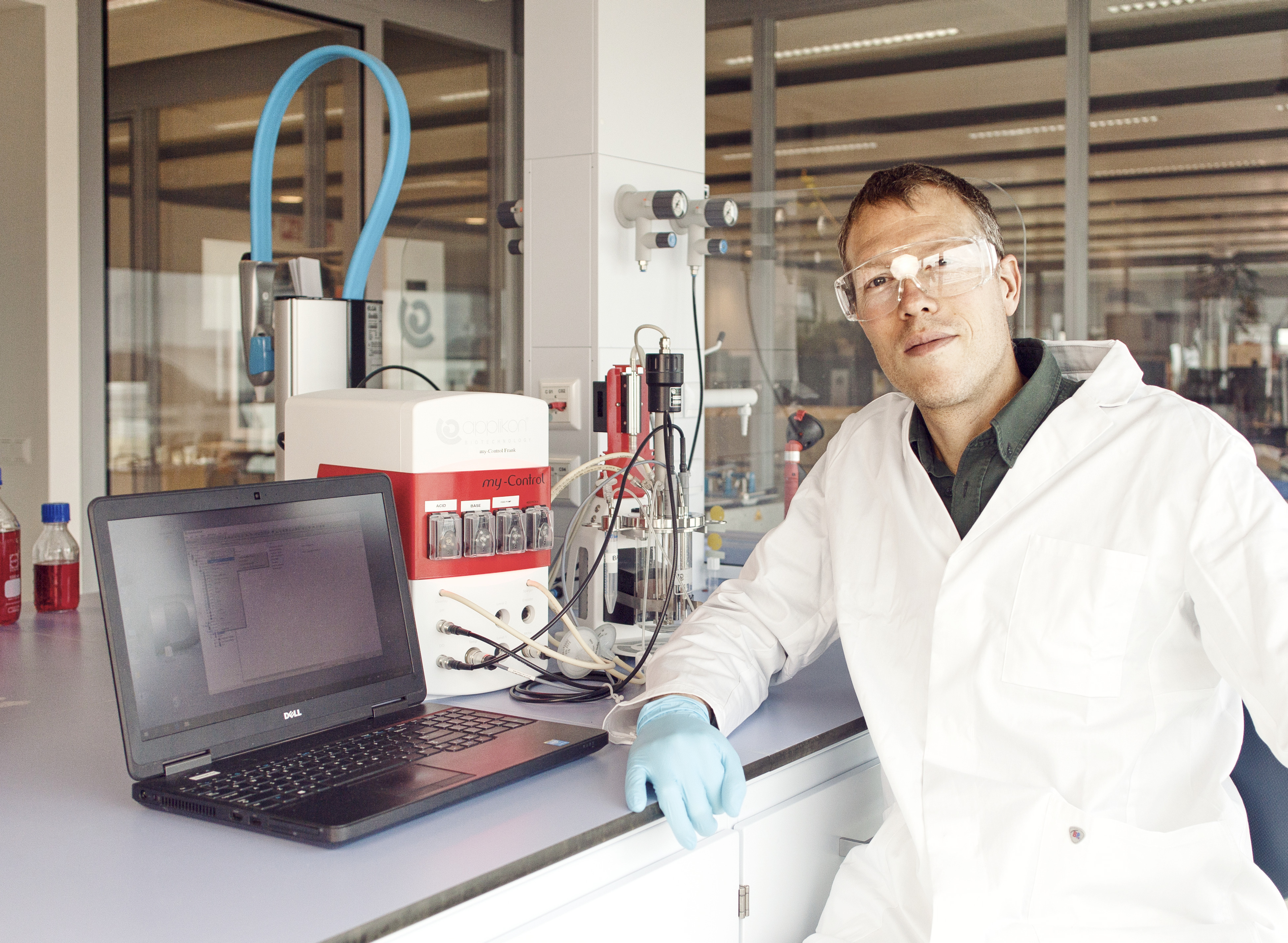 A person in a lab coat and safety glasses sits by a laptop and scientific equipment in a laboratory setting.