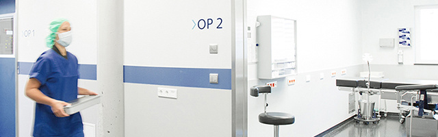Eine Person im OP-Kittel und mit Maske geht mit einem Tablett an einem Operationssaal mit der Aufschrift „OP 2“ in einem Krankenhausflur vorbei.