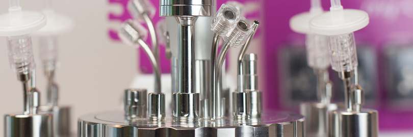 Nahaufnahme eines glänzenden, metallischen Laborinstruments mit verschiedenen Schläuchen und Anschlüssen vor einem violetten Hintergrund.