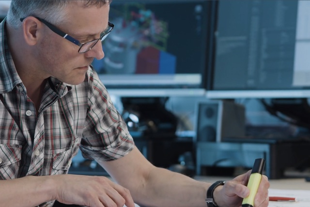 Un homme en chemise à carreaux et lunettes écrivant à un bureau avec des écrans d'ordinateur en arrière-plan.