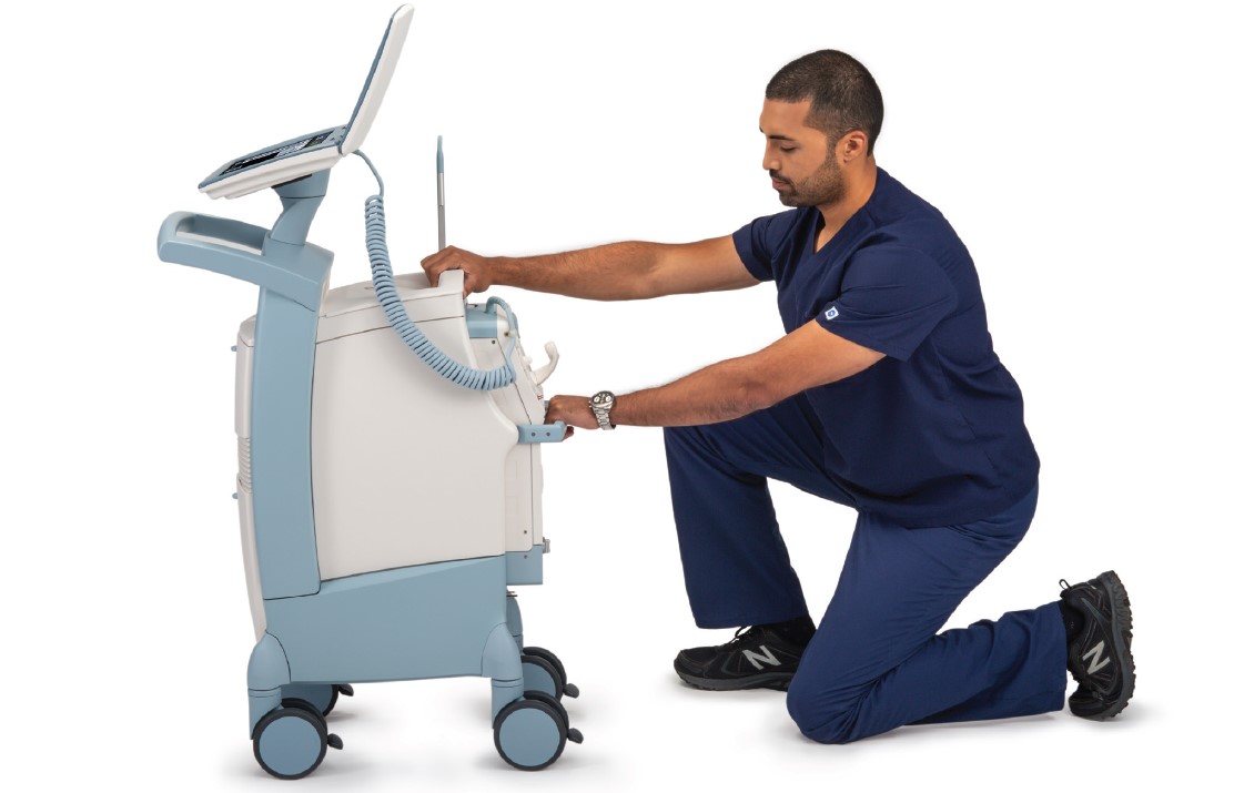 Person in blue scrubs kneeling, adjusting a medical machine on wheels in a clinical setting.