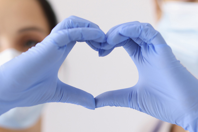 Two people wearing masks and blue gloves form a heart shape with their hands.