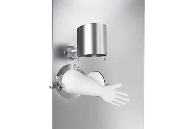 Sterile glove attached to metallic wall in cleanroom setting, with cylindrical metal container above.