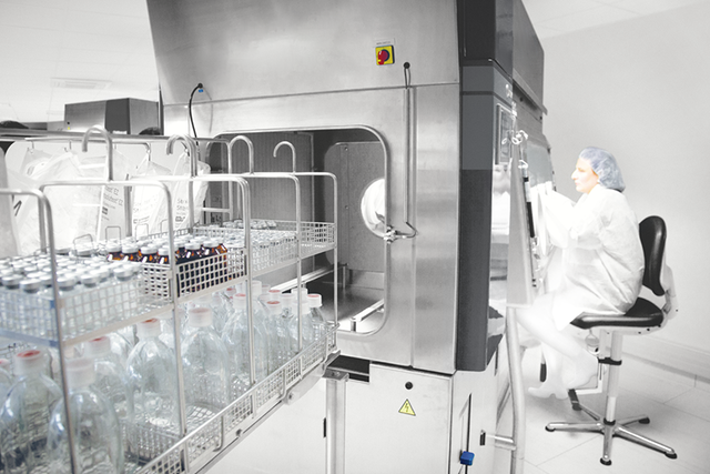 Opérateur en combinaison adaptée aux environnements stériles travaille sur un isolateur Isotest de ww5. Au premier plan, des composants et des dispositifs prêts pour le test de stérilité.