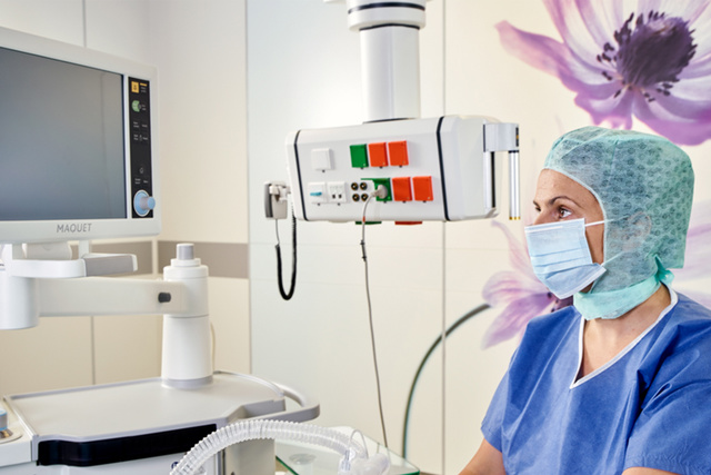 Medical professional in scrubs and mask, sitting near medical equipment, with a floral wall design in the background.