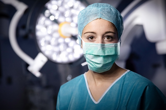 A healthcare worker in scrubs and a mask stands in an operating room, with surgical lights in the background.