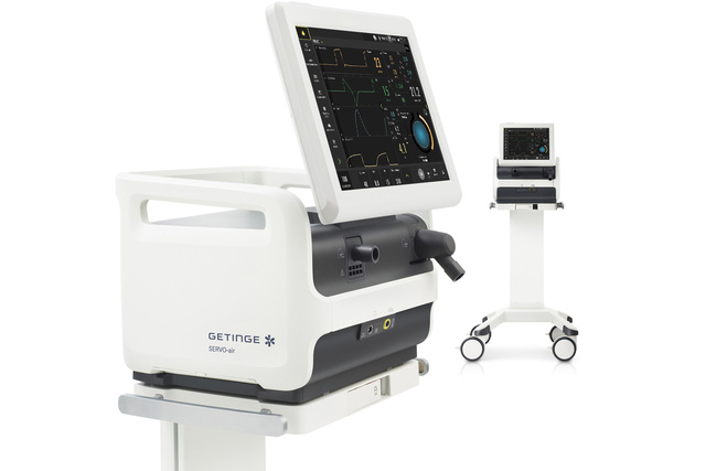 Two medical ventilators, each with a display screen showing graphs and data, on white wheeled stands.
