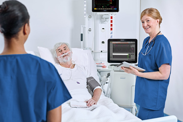 Image d'un patient âgé allongé dans un lit d'hôpital relié à un Servo-u et un bras de distribution, assisté de deux infirmières.