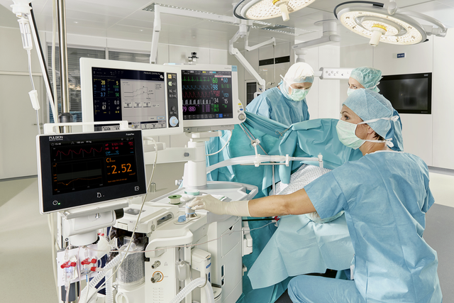 Professionnels de santé en tenue chirurgicale opérant dans un bloc opératoire et regardant des écrans PulsioFlex montés sur une station d'anesthésie Flow-i