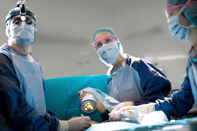 Surgeons in scrubs and masks perform a medical procedure under bright lights in an operating room.