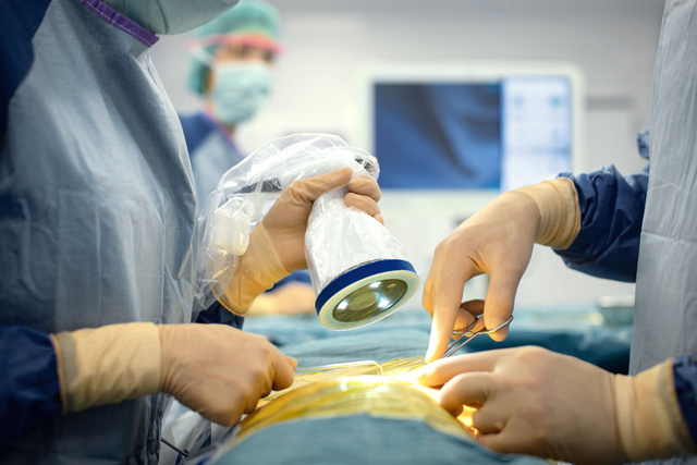 Surgeons with gloves and masks perform surgery, using medical instruments and a light device.
