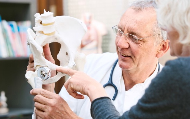 Doctor explaining hip joint model to a patient in an office filled with books.