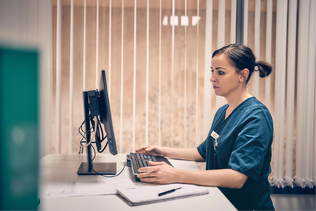 Healthcare worker in scrubs typing on a computer at a desk with a notebook, in front of vertical blinds.