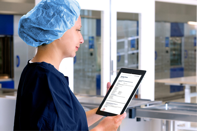 A healthcare worker in scrubs and a hairnet looks at a document on a tablet in a clinical setting.