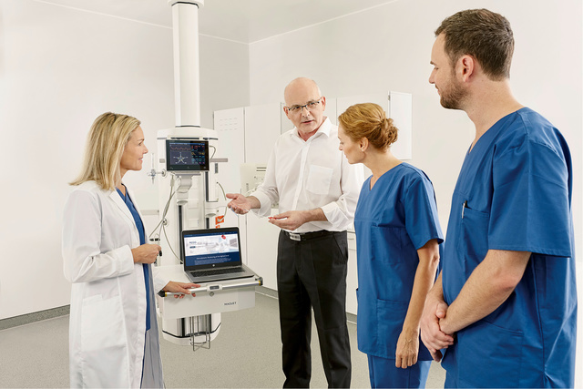 Medical team discussing next to a medical device and a laptop, with two in blue scrubs and one in a white coat.