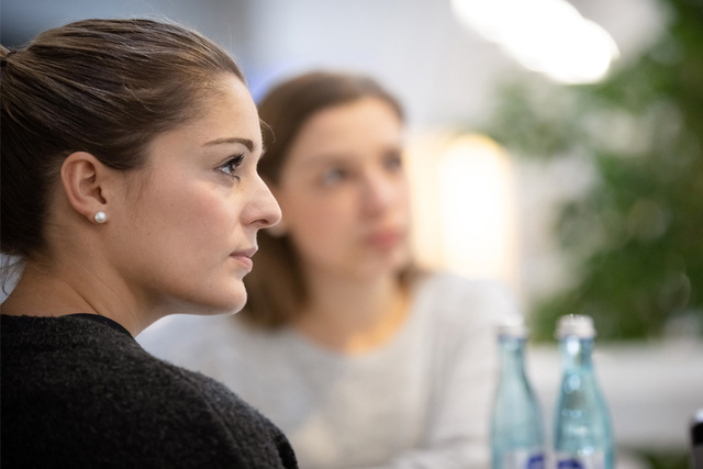 Zwei Frauen sitzen und hören aufmerksam zu, im Hintergrund unscharfes Grün und Wasserflaschen auf dem Tisch.