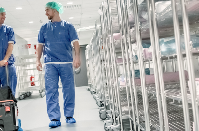 Two healthcare workers in blue scrubs and green caps walk through a hospital corridor, pulling a wheeled equipment case.